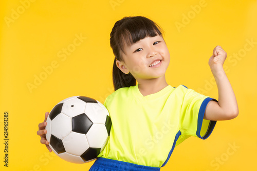 Foto A high-energy portrait of the same East Asian girl on a solid yellow backdrop, holding the soccer ball aloft with one hand while cheering excitedly and raising her other fist in a victorious gesture