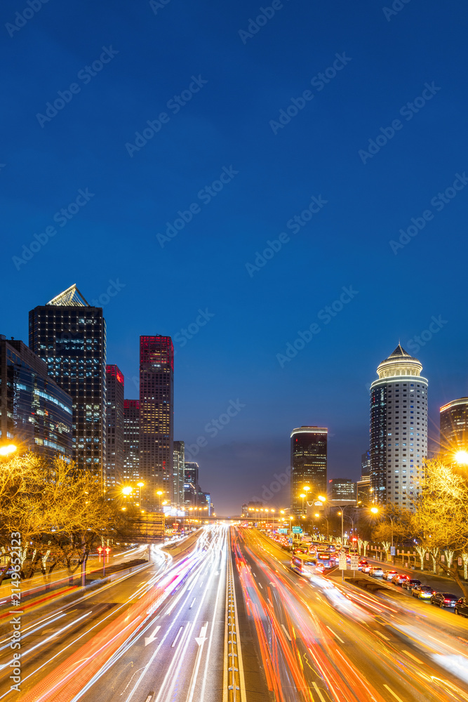 Obraz premium A long-exposure shot of an urban highway at night, showing light trails from moving vehicles flanked by illuminated skyscrapers under a dark blue sky.