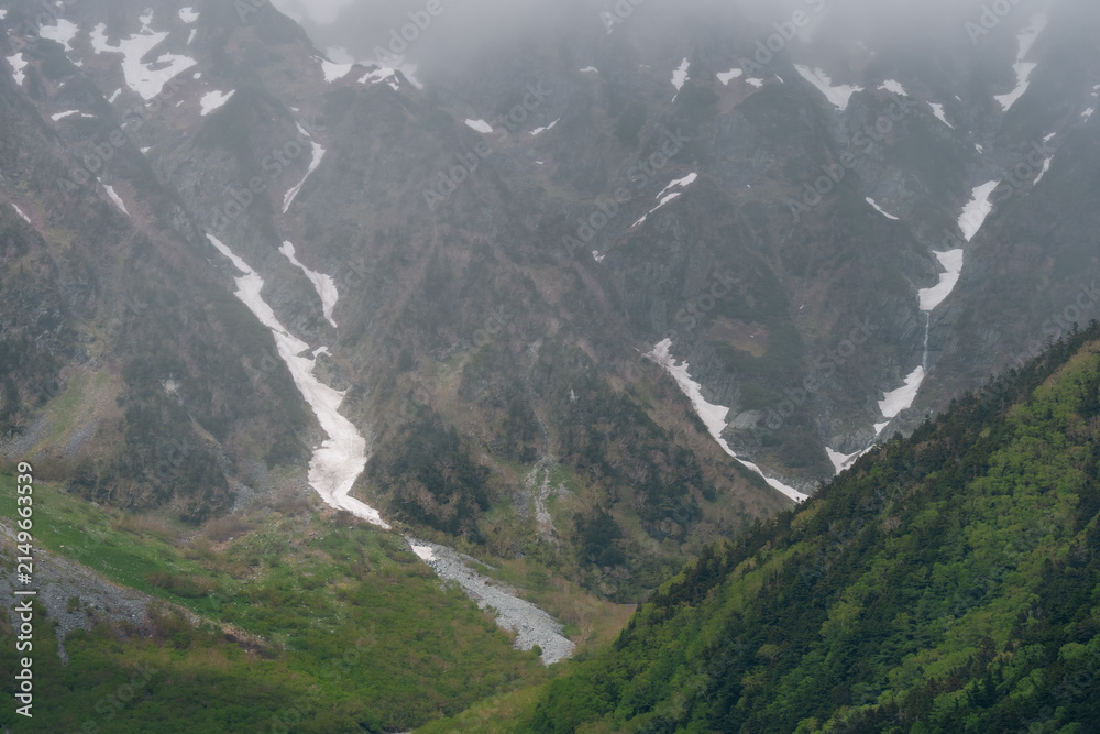 Obraz premium Mountain valley snow forest alpine peak mist cloud rocky slope green hill river landscape with rugged terrain, foggy sky, glacier patch, wilderness, and remote nature scenery