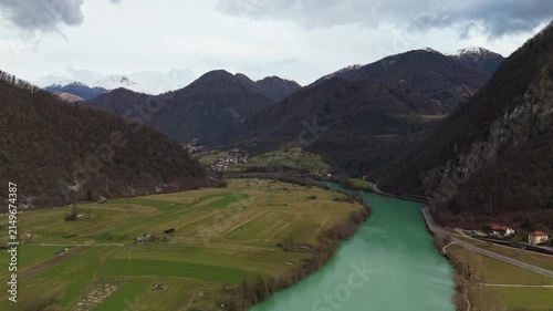Most na Soči, Slovenia, with the emerald Soča River winding through a forested alpine valley beneath snowy Julian Alps peaks, captured by drone above rural fields and roads.