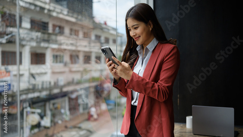 Smiling businesswoman using a smartphone while standing beside a large window in a modern office or café.