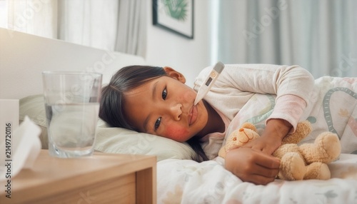 Sick child lying in bed with blanket, thermometer in mouth.