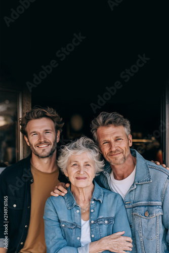 Smiling Multigenerational Group Portrait of Two Men Embracing an Elderly Woman in a Denim Jacket against dark background, Celebrating Family Love, Togetherness and Connection. Copy Space, 2:3.