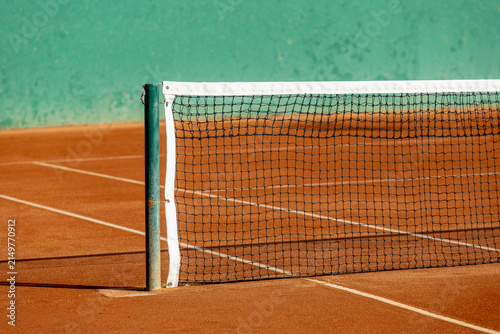 Close-up of a tennis net on a red clay court with white boundary lines and a green background wall under bright sunlight