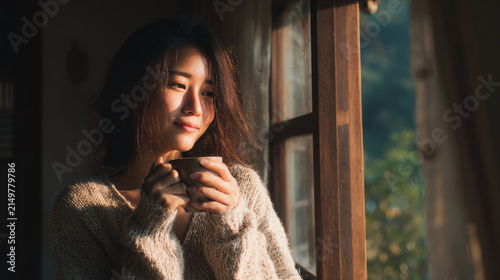 Thoughtful young woman savoring a hot beverage while sitting by the window bathed in gentle natural light