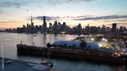 High-angle aerial shot circling the long steel hull, self-unloading boom, and superstructure of a cargo ship moving slowly through the harbor channel, revealing the glowing Toronto skyline at dusk