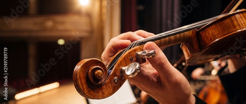 Violin scroll and tuning pegs crafted from polished maple wood held by musician during orchestral performance in dim vintage concert hall