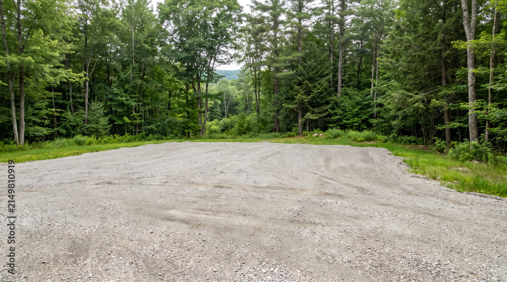 Obraz premium Empty gravel lot land, with cleared ground and tire tracks, extending towards green forest trees, under overcast sky in rural area, real estate land photography