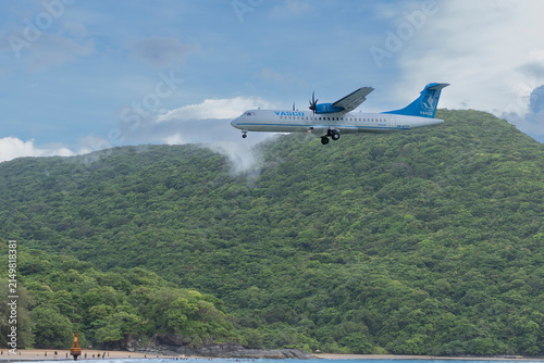 A VASCO turboprop airplane flies low over the ocean on final approach to land at Dam Trau Beach in Con Dao, Vietnam, with a tropical green mountain in the background.