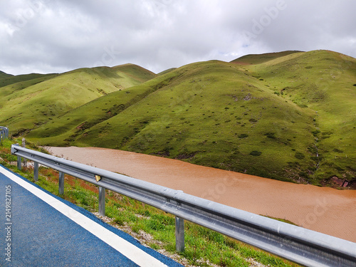 A scenic mountain road with a metal guardrail running alongside a river and lush green alpine grasslands at Dahai Caoshan (Dahai Grassland) in Huize, Qujing, Yunnan, China, under a cloudy sky.