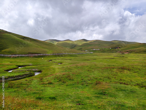 Wide angle view of the vast green alpine meadow with a white wooden fence stretching along rolling hills at Xiao Xiniu (Small Rhinoceros) viewpoint in Dahai Caoshan, Huize, Qujing, Yunnan, China, unde