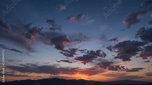 A wide shot of a sunset over a distant mountain range, where the sky is layered with clouds shifting from dark blue and purple at the top to vibrant orange and red near the horizon, 4K, HD Video.