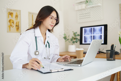 Female doctor reviewing medical records on a clipboard while working with a laptop in a bright modern clinic.