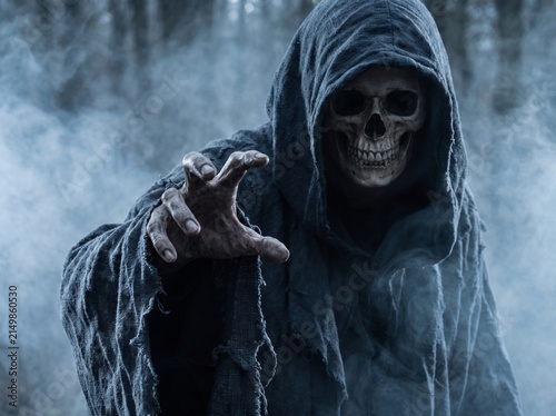 Grim reaper wearing dark hooded robes reaches out with bony hands amid thick eerie fog in a dark mysterious forest setting