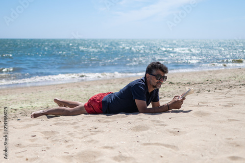Obraz na plátne Man lying on the sand reading a book, enjoying a peaceful summer vacation by the