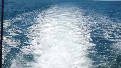 Wide rear view from fast moving ship stern showing powerful white foam wake trail on deep blue ocean surface. High speed maritime vessel navigation creates strong turbulent splash and spray effect. 4k