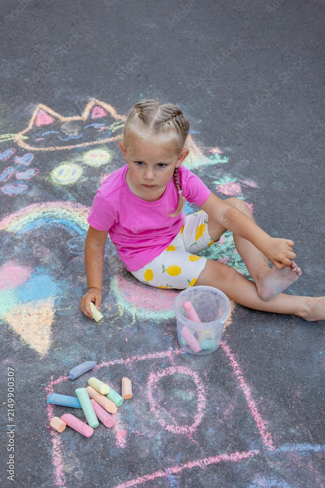 Naklejka premium Little girl draws with colorful chalk on the asphalt on the street in summer