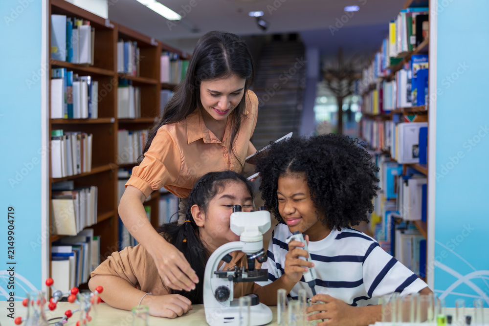 Obraz premium A female teacher guides two diverse young female students as they conduct an engaging science experiment using a microscope and chemistry beakers in a school library.