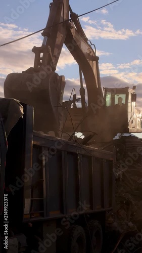 Heavy construction excavator loading earth and mud into a large dump truck at sunset on a worksite.