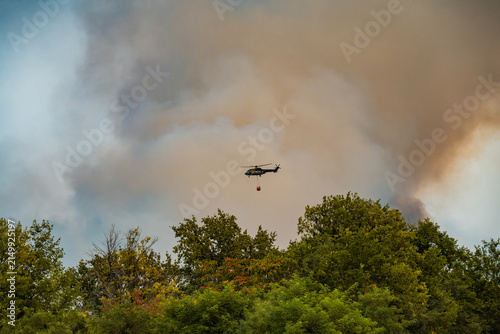 Firefighting Airbus H215 Super Puma helicopter flying with a Bambi bucket over a country road into dense wildfire smoke in Deliblatska Pescara. Deliblato, Serbia - August 7, 2026.
