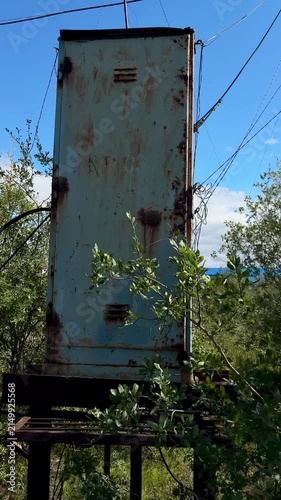 An abandoned, weathered blue metal structure with heavy rust, standing amidst green foliage and bushes under a clear blue sky. Industrial decay in nature.