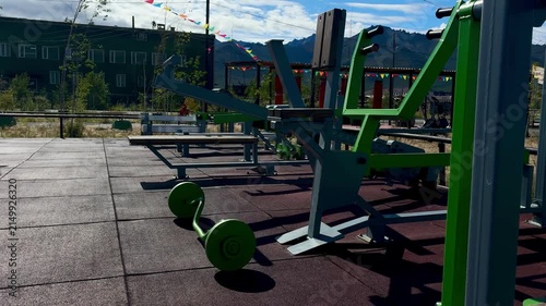 A modern green weightlifting machine at an outdoor fitness station in a scenic park with mountains and colorful bunting in the background.