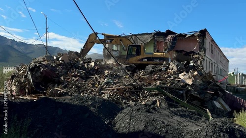 A heavy excavator machine working amidst a massive pile of construction debris and rubble near mountains under a blue sky.