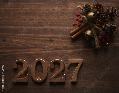 Golden numbers 2027 are set against a rich wooden background, adorned with rustic holiday decorations featuring pinecones, red berries, and cinnamon