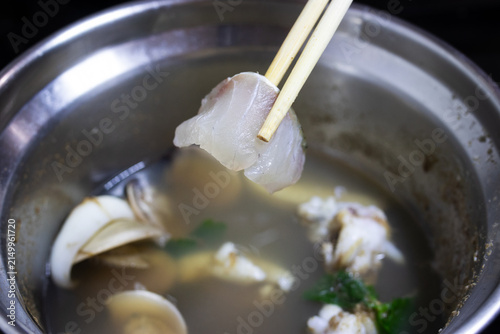 Chopsticks holding raw fresh fish slice over boiling hotpot soup in stainless pot.
