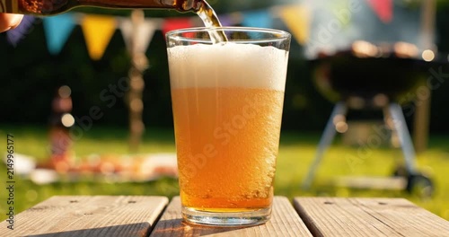 Beer pouring from bottle into glass on wooden table outdoors, summer garden party barbecue setting.
