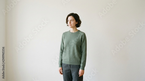 Young woman in a green sweater and grey pants stands against a white wall, showcasing casual, modern style, representative of everyday fashion.