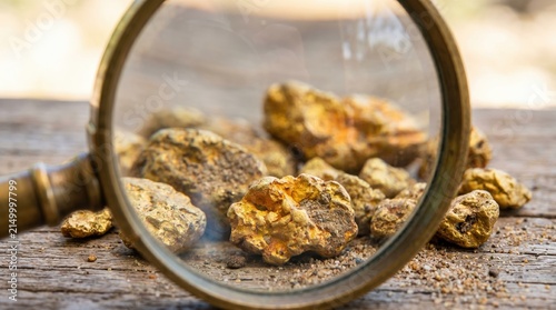 Gold nuggets under a magnifying glass on a rustic wooden table representing mineral prospecting, precious metal discovery, and wealth accumulation