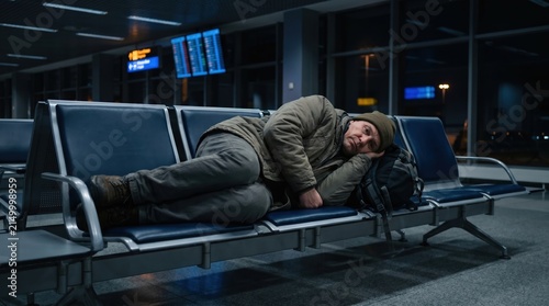 Middle-aged caucasian man sleeping on airport waiting room chairs with backpack waiting for delayed flight travel disturbance concept