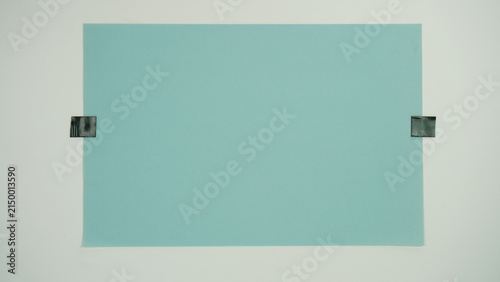 Bild auf Leinwand A piece of blue paper positioned horizontal and attached to a surface with black tape on two sides, isolated on a white background