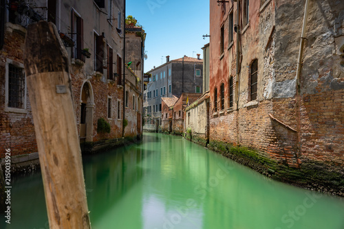 Venice canal with green water and historic brick buildings in Italy bathed in bright sunny summer light under clear blue sky