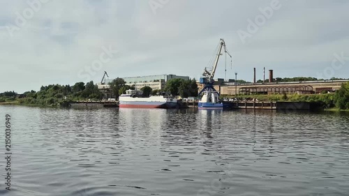 Cargo ship docked at industrial river port. Industrial infrastructure near waterway. Summer river cruise view of logistics harbor with crane and factory building.