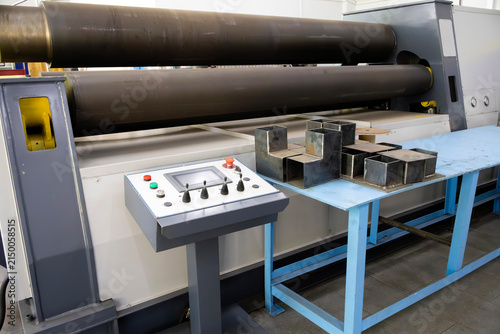 Heavy-duty four-roll bending machine forms metal plates; industrial control panel enables precise operation. Steel profile samples on blue workbench highlight fabrication workflow in a production sett