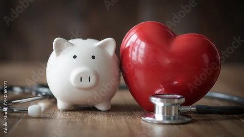 Stethoscope resting on wooden desk next to red heart shape and piggy bank symbolizing healthcare savings