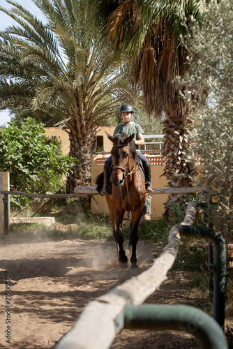 Young equestrian riding a brown horse in an outdoor riding arena, wearing a protective helmet and equestrian gear. Horseback riding lesson in a natural setting surrounded by trees, paddocks