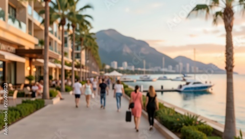 blurred view of luxury resort promenade at sunset with tourists, palm trees, marina with yachts, and mountain backdrop.