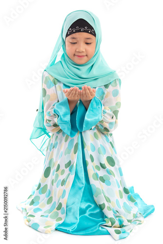 Charming young Asian Muslim child in traditional headscarf kneeling politely holding hands up in prayer looking down.