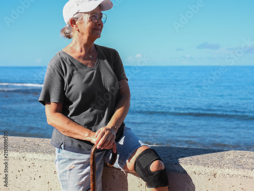 Bild auf Leinwand Senior woman with a knee brace and cane looking at the sea, embracing aging grac