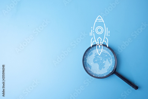 Global business startup and innovation concept. Magnifying glass focusing on a digital wireframe globe with a white rocket ship launching on a blue background with copy space.