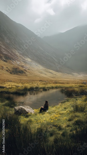 Person sitting by a placid stream in a valley nestled amongst mountains.