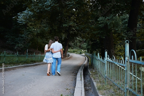 Konstfotografi Young couple walk arm in arm along a tree-lined park pathway, enjoying a quiet romantic summer stroll surrounded by green foliage, an old metal fence and a gently curving paved walkway