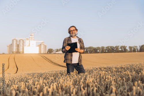 Obraz na płótnie Agronomist inspecting wheat crop in a field, holding a clipboard, grain elevator