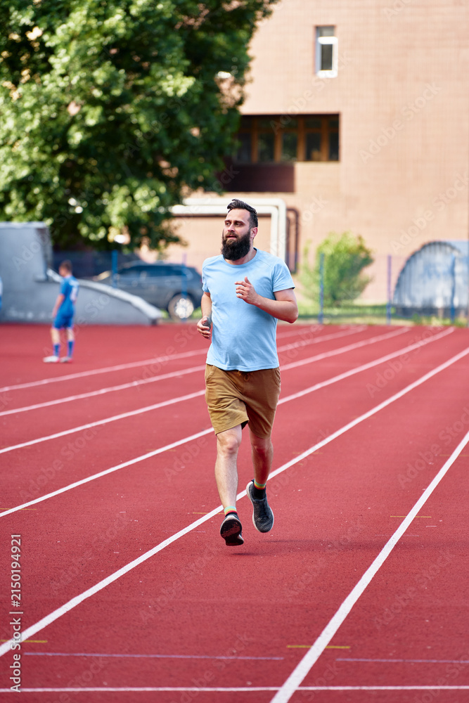 Obraz premium A man in casual clothing jogging on a red tar track outdoors during a sunny day, capturing concepts of exercise, fitness, and leisure in an urban environment.