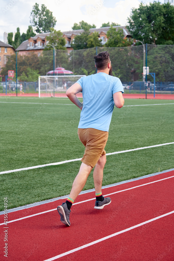 Obraz premium A man in casual attire jogging along a red athletic track in an outdoor sports environment with a green grass field and residential buildings in the background.