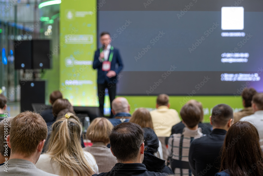 Fototapeta premium Speaker addresses a seated audience beside a dark presentation slide on a large screen.