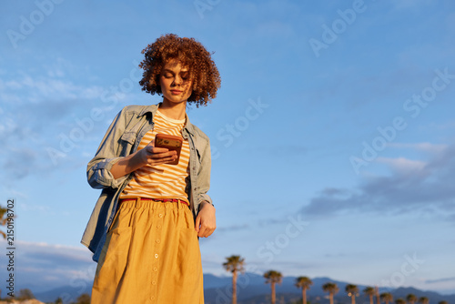 Young woman with curly hair stands outdoors in sunny weather, looking at her smartphone, wearing a striped top, denim jacket and mustard skirt, palm trees line the background.